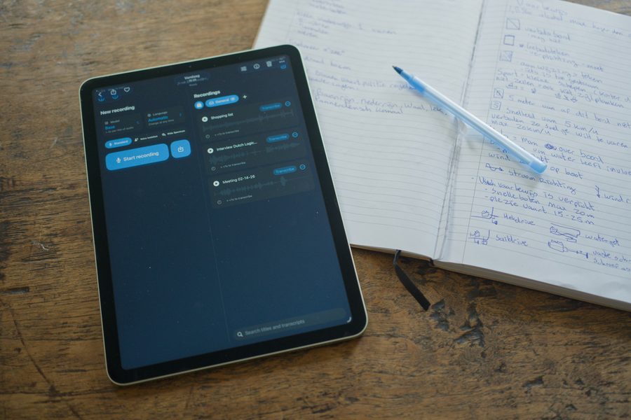 Een iPad in de donkere modus met de opnamelijst van Just Transcribe, naast een opengeslagen notitieboek en een pen.