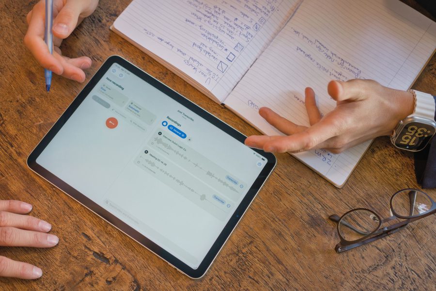 Een iPad op een houten tafel met Just Transcribe in de lichte modus, naast een opengeslagen notitieboek met handgeschreven aantekeningen.