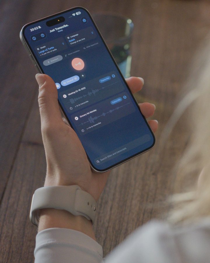 Een hand met een iPhone waarop Just Transcribe in de donkere modus staat, met Nederlands als taal.